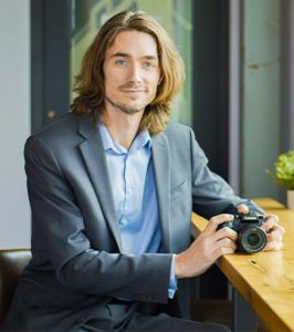 Image of Videographer/Editor Evan Castor in gray sport coat standing and holding a camera at Paradigm Digital Group offices