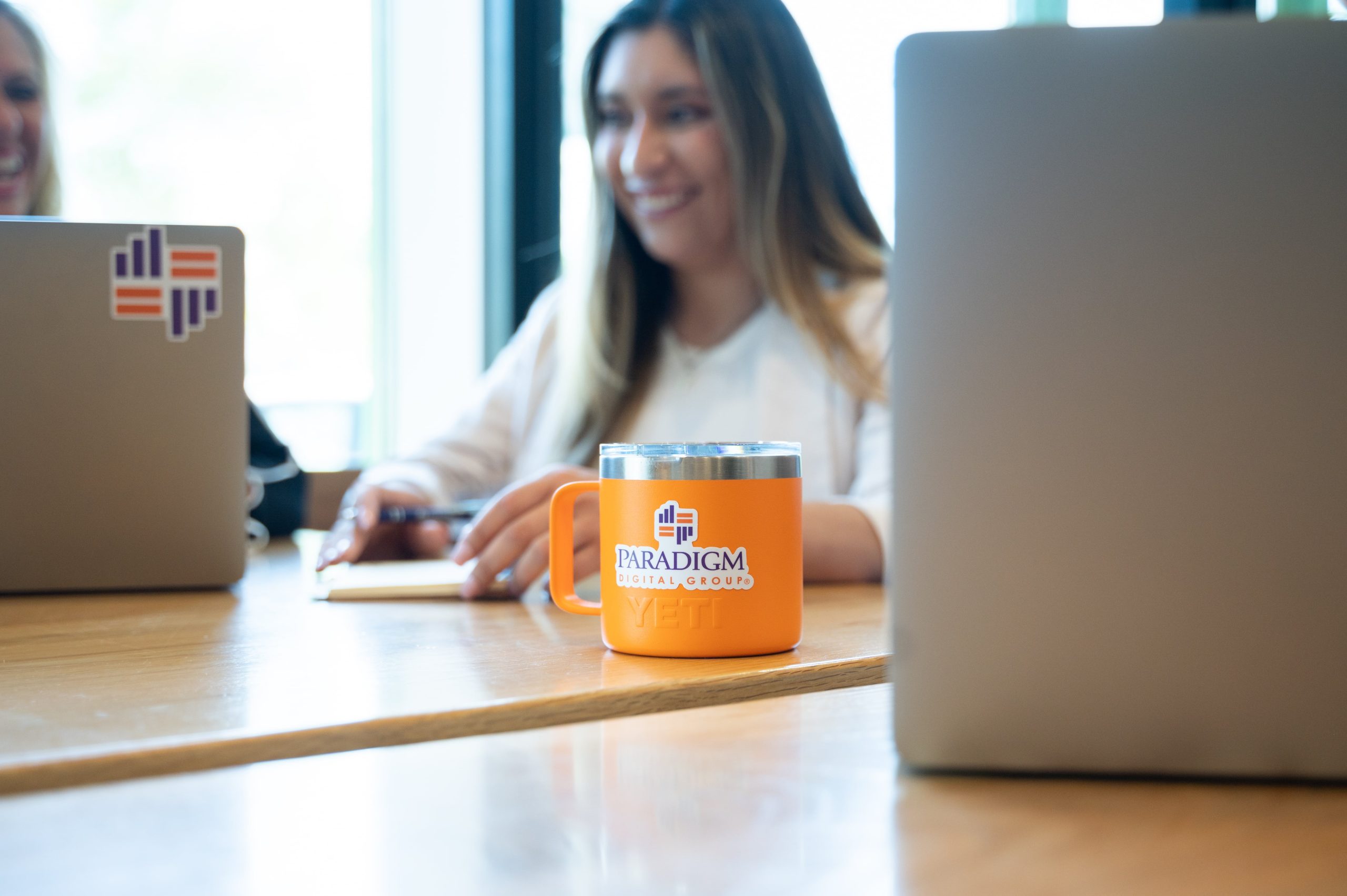 Image of a Paradigm Digital Group orange metal mug sitting on a table and a woman sitting in the background