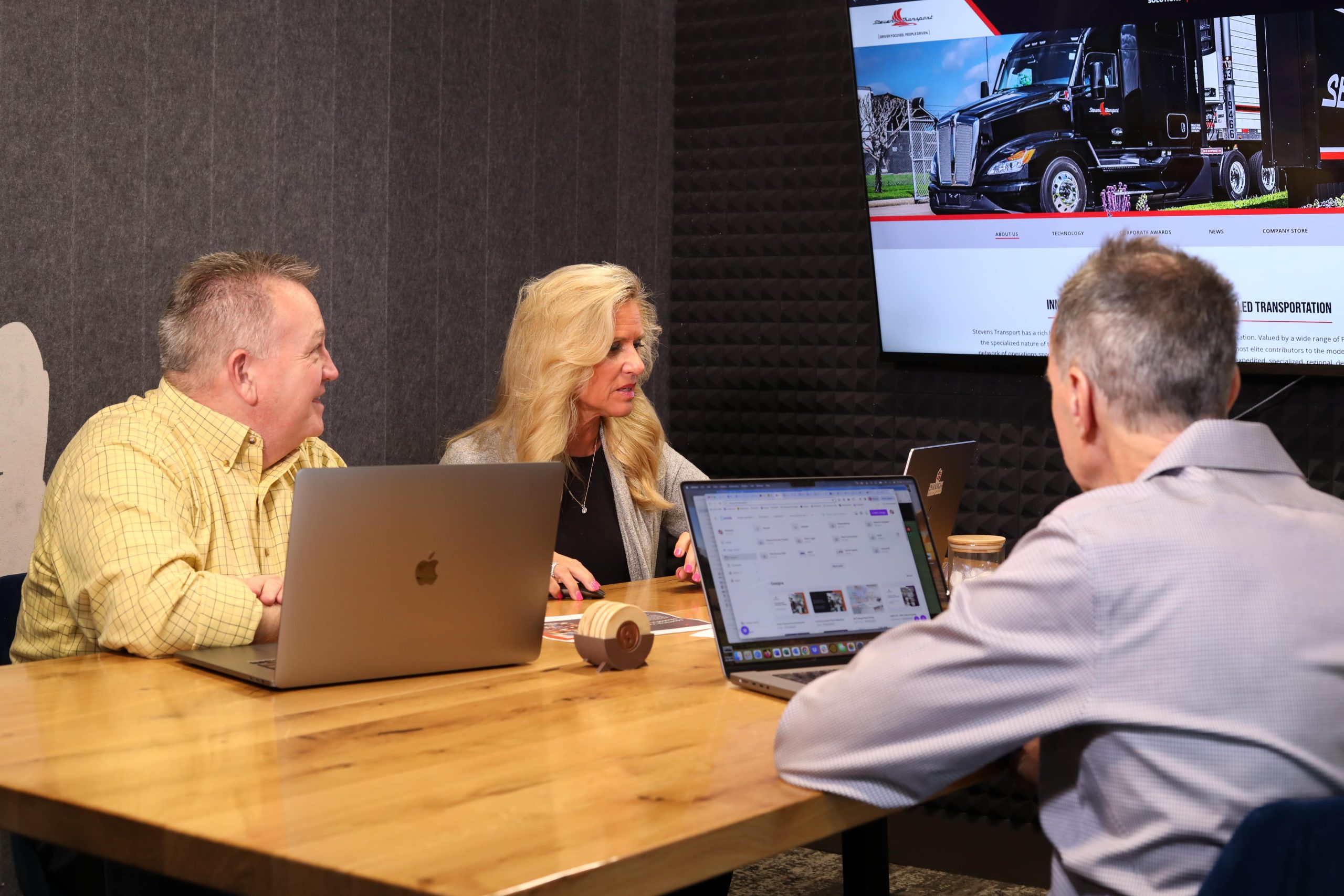 Bob, Deb, and Mike sitting at a table with laptops and looking at TV